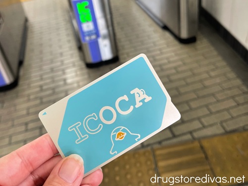 Fingers holding a blue ICOCA travel card in a Japanese train station.