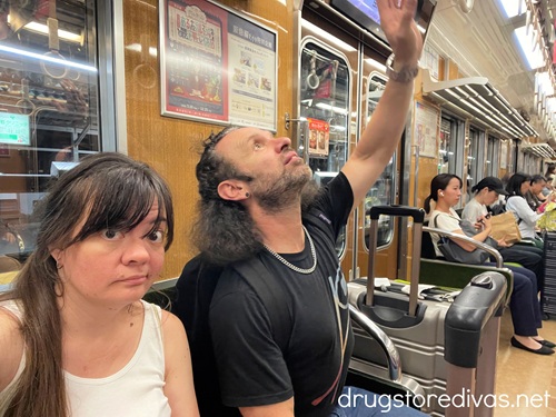 A man and woman sitting on a subway in Japan with the man reaching up.