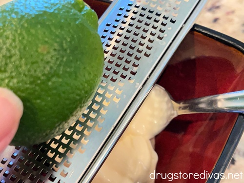 A lime on a microplane grater above a maroon bowl with mayonnaise in it.
