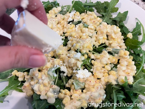A hand crumbling white cheese on top of corn and arugula. 
