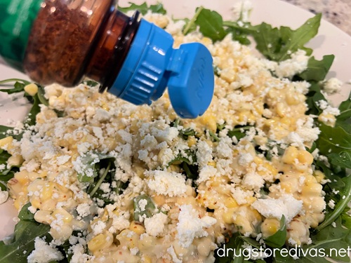 Tajin seasoning in a bottle being sprinkled over corn kernels and arugula in a bowl.