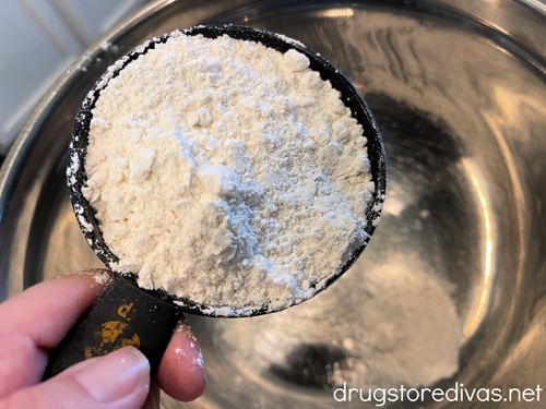 A hand holding a measuring cup of flour above a silver bowl.