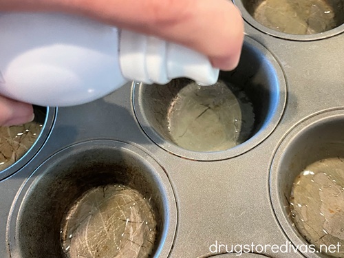 A hand spraying cooking spray in cavities of a muffin pan.