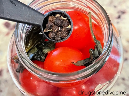 A measuring spoon of black peppercorns above a jar with cherry tomatoes and basil in it.