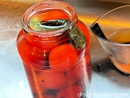 A glass jar filled with cherry tomatoes, basil, and a liquid in the sink with an empty measuring cup behind it.