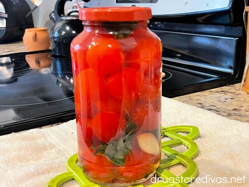 A glass jar with cherry tomatoes, basil, and garlic on it on top of a green trivet and white towel near a stove.