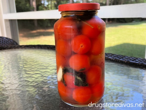 A glass jar with a red top filled with cherry tomatoes, basil, and garlic on a glass table.