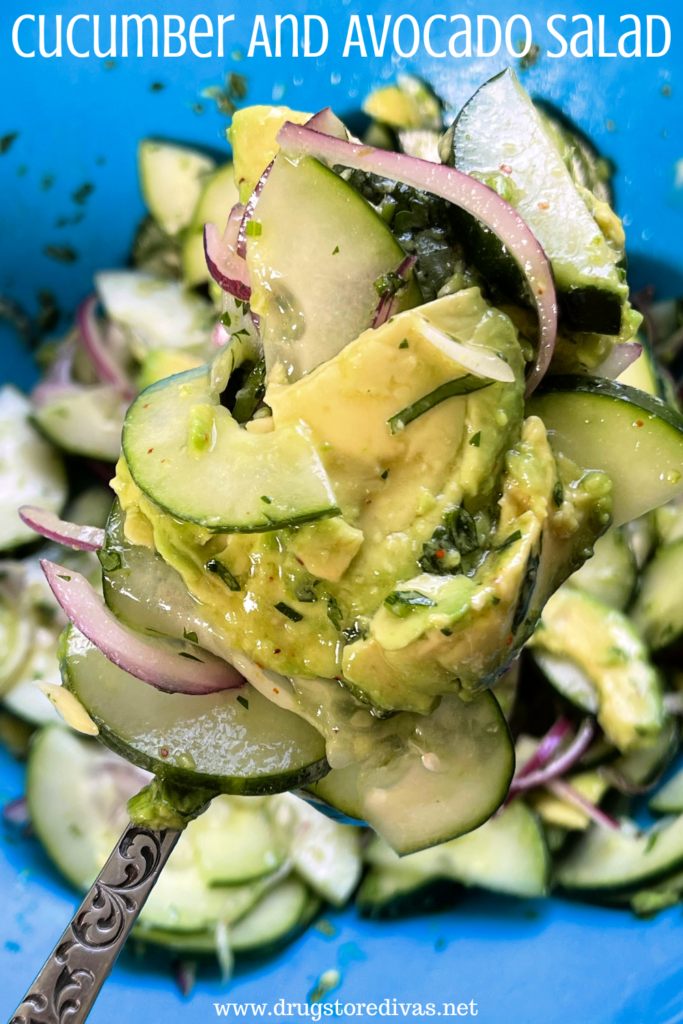 A spoon holding a scoop of salad with avocado, cucumber, and red onion it in above a blue bowl with the words "Cucumber And Avocado Salad" digitally written on top.