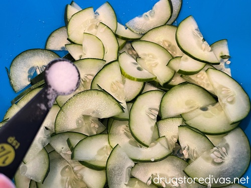 Salt in a measuring spoon being poured on cucumber pieces in a blue bowl.