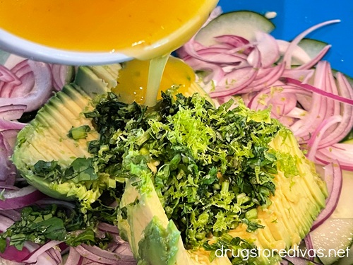 An orange salad dressing in a light blue bowl being poured over cilantro, avocado, red onion, and cucumber in a blue bowl.
