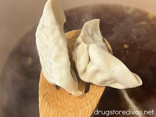 Two gyoza in a wooden spoon above a Dutch oven with brown liquid in it.