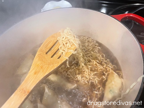 A wooden spoon holding up instant ramen noodles above a Dutch oven with brown liquid and gyoza in it.