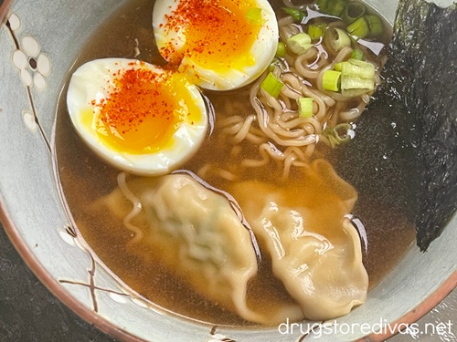 A jammy egg cut open with toharashi on top in a bowl of broth with two gyoza, ramen noodles, green onion, and a piece of seaweed.