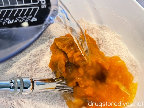 Water in a measuring cup being poured into a white bowl with flour and canned pumpkin in it.