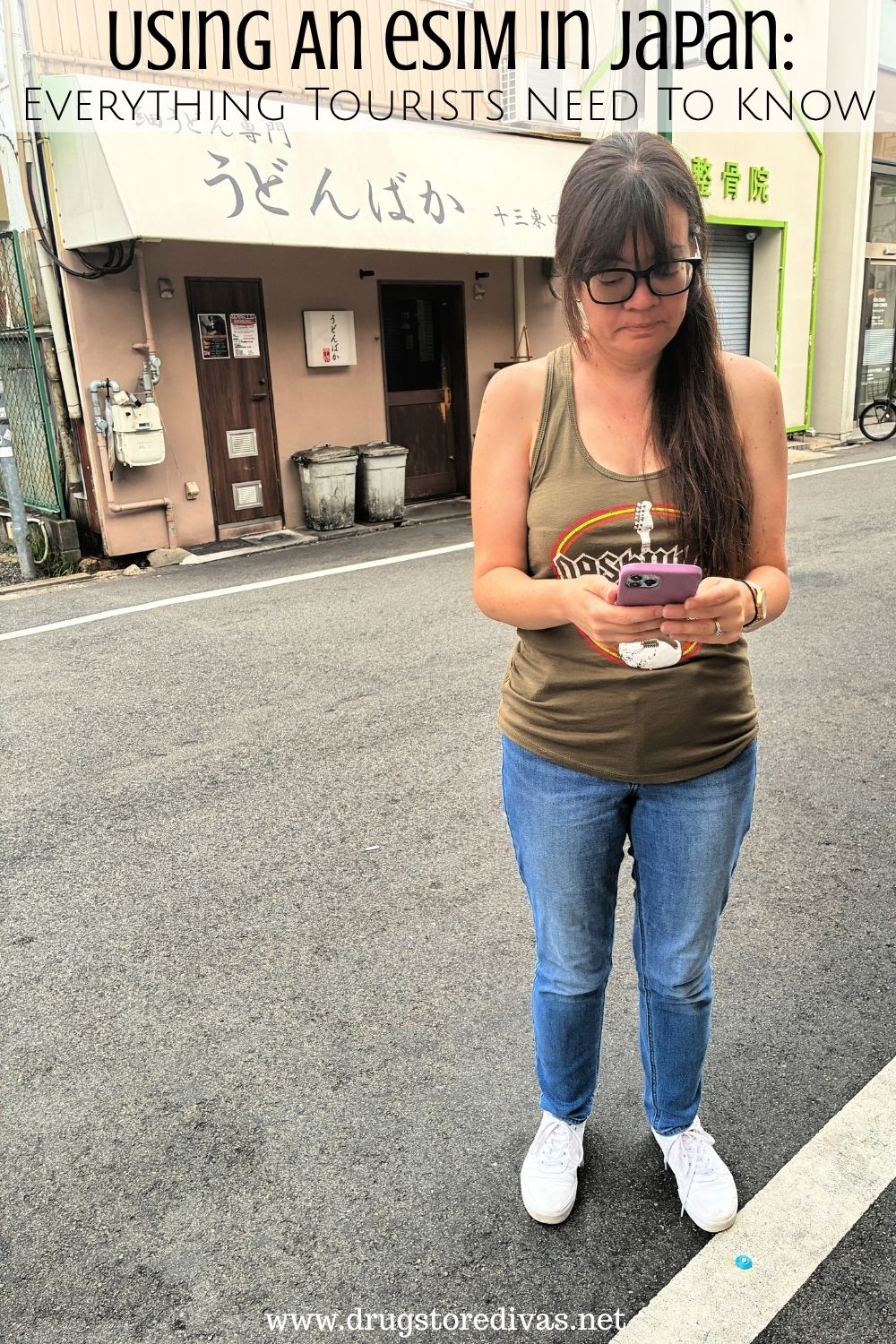 A woman in jeans and a tank top, looking at her phone, on a Japanese street with the words 